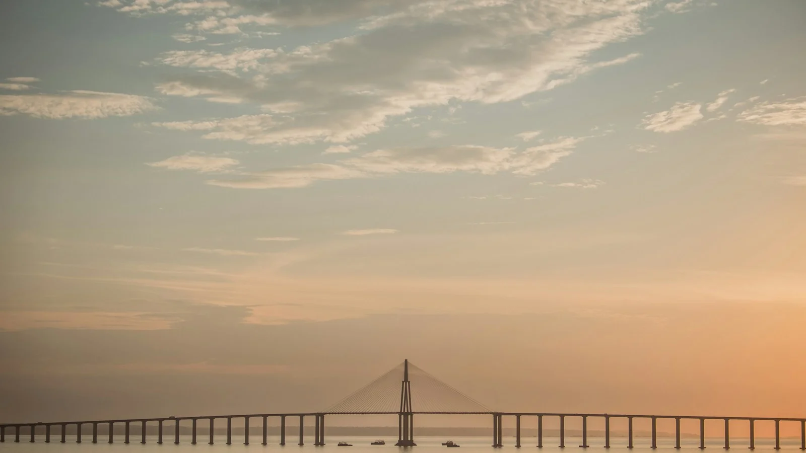 A stunning photograph captures the immense scale of the Rio Negro Bridge in Manaus, Brazil, as it stretches across the water under a soft, pastel-hued sky. The cable-stayed mid-section of the bridge creates a striking focal point, silhouetted against the warm tones of either sunrise or sunset. The tranquil water reflects the gentle light, and a few small boats can be seen, adding a sense of life and scale to the serene and majestic scene. This image perfectly encapsulates themes of modern architecture, travel, and the peaceful beauty of the Amazonian landscape.